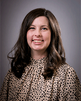 A woman with long brown hair smiling with a black polka dotted shirt. 