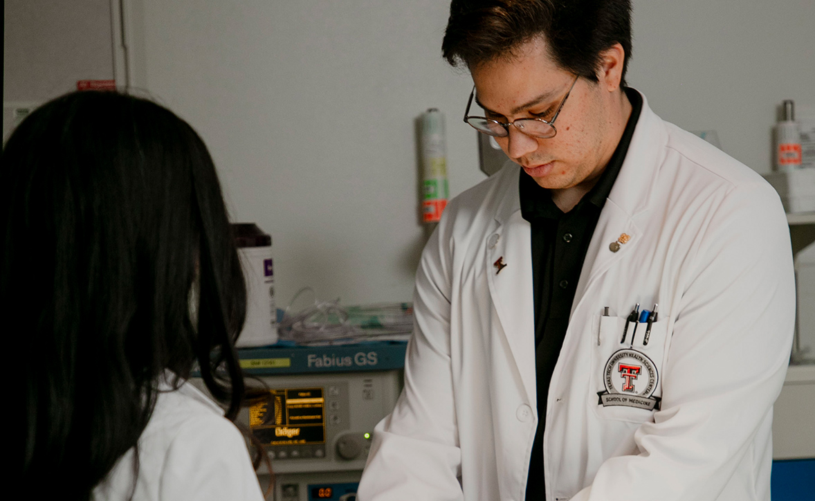 Two medical professionals in white coats, one with a Texas Tech University Health Sciences Center School of Medicine badge, working together in a clinical setting with medical equipment in the background.