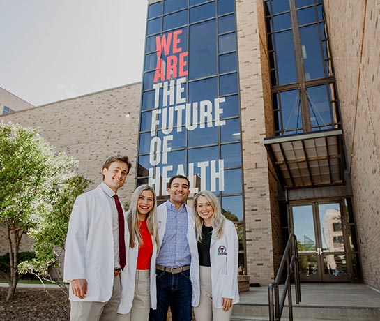 Four students standing infront of a building with we are the future of health sign behind them Four students standing infront of a building with we are the future of health sign behind them