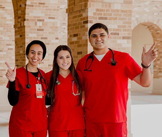 Three students in red scrubs standing next to each other smiling with tech guns up Three students in red scrubs standing next to each other smiling with tech guns up